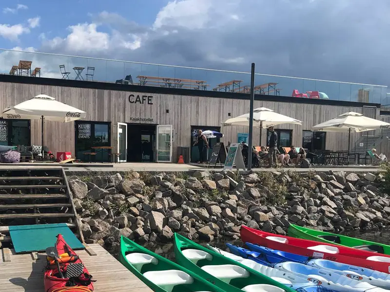 Kajakker i vandkanten og en rød på broen. Der er en trappe op mod Kajakhotellet hvor parasollerne er slået op man kan se borde og stole på tagterrassen.