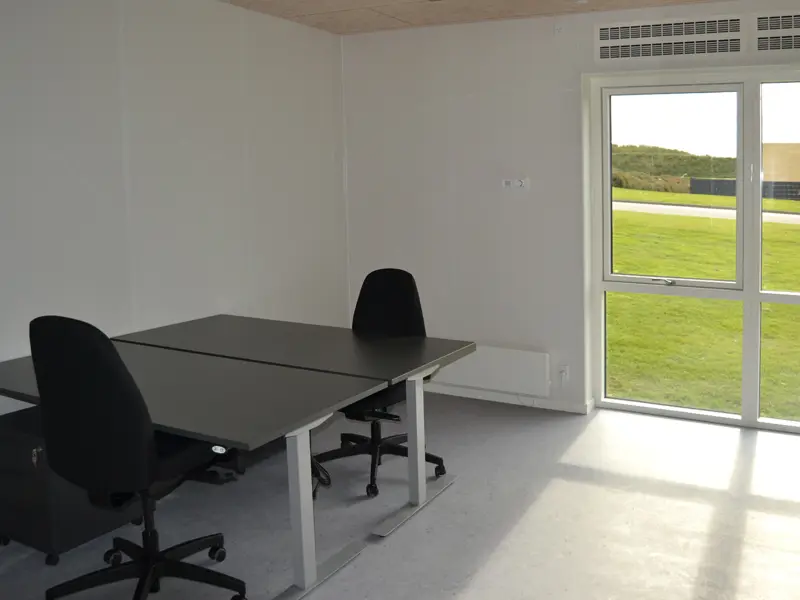 2 sorte kontorborde med kontorstole, placeret i et mindre kontorlokale.