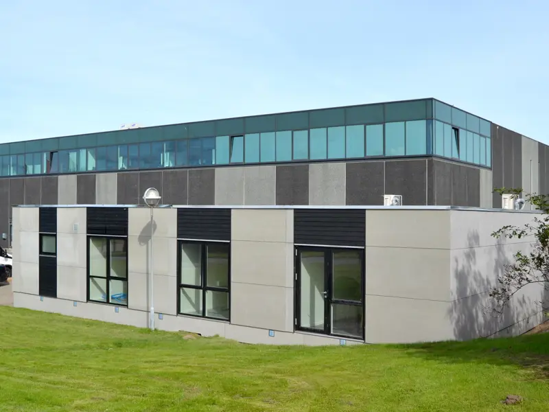Kontorbygning, bygget til eksisterende bygning. Grå facade og sort træbeklædning over vinduerne, der er græs foran bygningen.