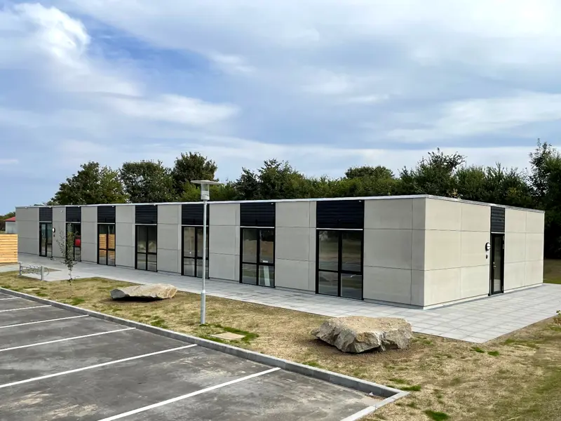 Bygning til hjemmeplejen Østermarie, der er plantet græs rundt om, lagt et par meget store sten og anlagt parkeringsplads.. Bygget i Flexicubes® med grå facader og træbeklædning over vinduer
