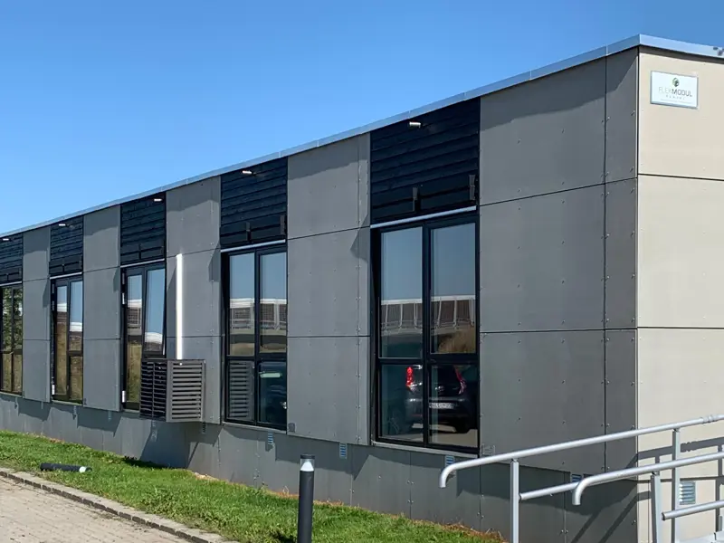 Grå bygning med træbeklædning over alle vinduer. Græs foran og man kan ane rampen op til indgangen.