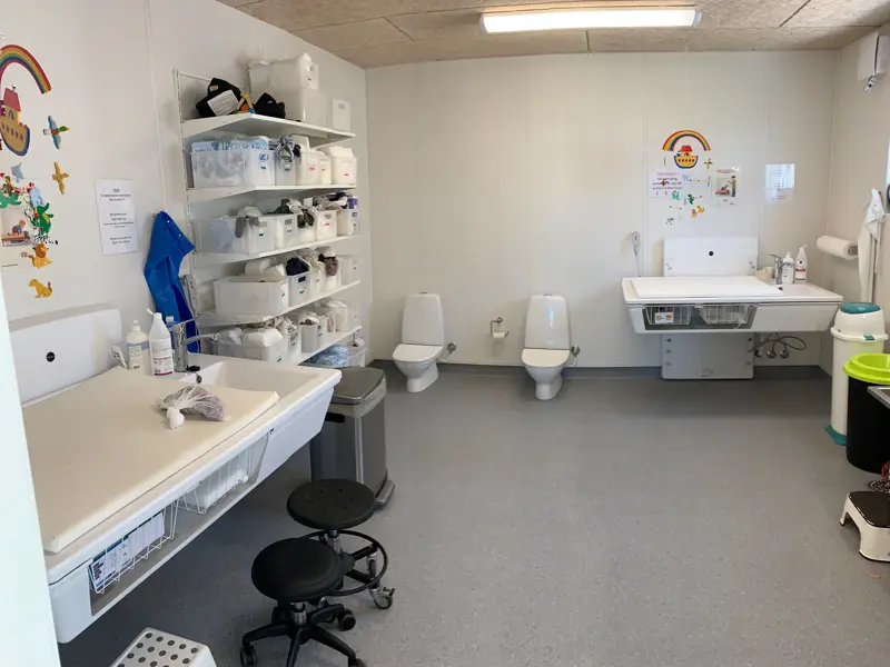 Toilet og puslerum i børneinstitution, en reol hvor hvert barn har en kasse med ekstra tøj og bleer. 2 hævesænke pusleborde og små børnetoiletter.