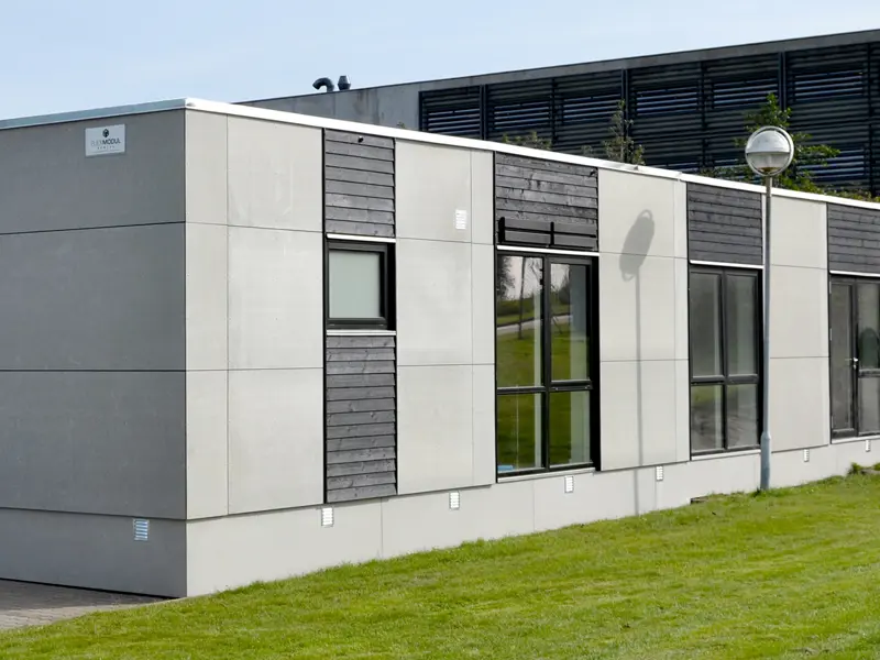 Grå bygning med træbeklædning ved vinduer. Solen skinner på bygningen og der er grøn græs foran.